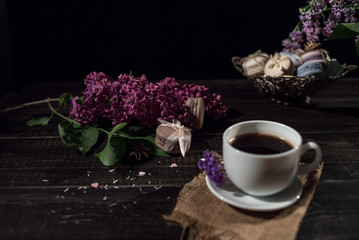 Obraz na płótnie Canvas Cup of coffee on linen napkin and several macarons in braid vase with branch of lilac.