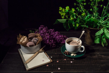 Fototapeta na wymiar Coffee and cream macaroons in a white plate with coffee cup, eco notebook with a pencil under lilac branch 