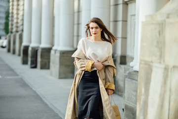 Slim elegant woman with red lips in a beige coat and black skirt brunette with long legs near the columns.