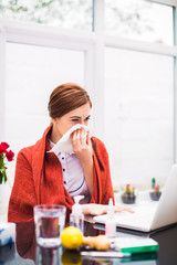 Portrait of young sick office worker who sits at the table with medicaments at the office, keeps napkins in hand and wipes nose