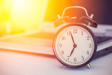 Old analog alarm clock and blurred laptop on white wooden table