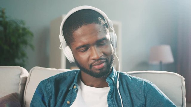 Attractive black african man listening to music in big white headphones while sitting on the couch at home. Slow motion. Close up