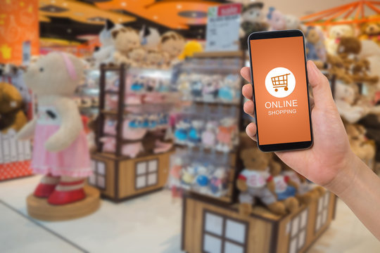 Human Hand Hold Smartphone, Tablet, Cell Phone With Online Shopping Application Screen On Blurry Toy Shop Background.