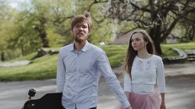 Young Couple In Love Walking In Summer Park Holding Hands. Woman In Pink Tutu Skirt Man With Longboard In Hands Three-axis Gimbal Stabilized Tracking Shot