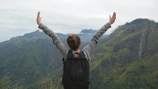 Woman tourist standing on edge of beautiful canyon, victoriously outstretching arms up and enjoying freedom. Young female hiker with backpack reaching up top of mountain and raised hands. Slow motion