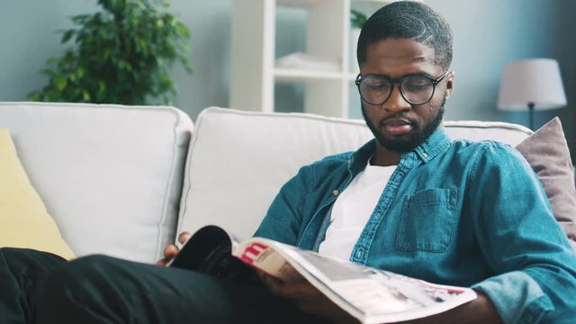 Young african man with beard in blue shirt sitting on white sofa and concentrated viewing magazine for shopping. Indoor.