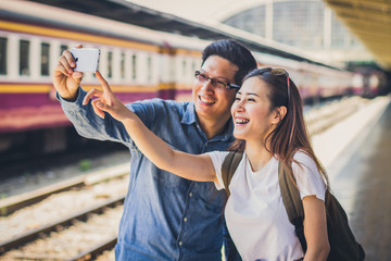 Happy Asian couple or friends reading on a phone while standing on train platform. Travel, holiday or commuting concept. Copy space.