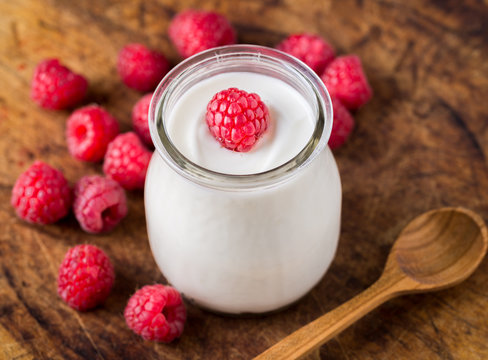 White yogurt with raspberries in glass bowl on rustic wooden table.
