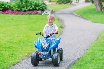 Portrait of smiling little Caucasian boy toddler driving blue electric car on the road path outside on spring summer day, seasonal child activity concept, healthy childhood lifestyle