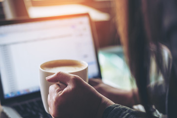 A business woman holding coffee cup while using and typing on laptop in office