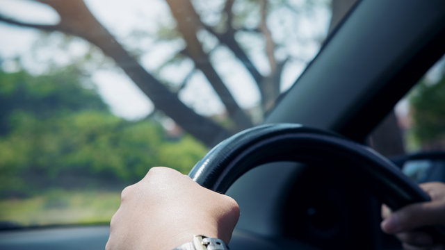 Woman's hands holding on black steering wheel while driving a car with big tree background