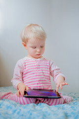Cute adorable white Caucasian blond baby girl sitting in bed playing with digital tablet with funny face expression, candid lifestyle, early development, new technology generation