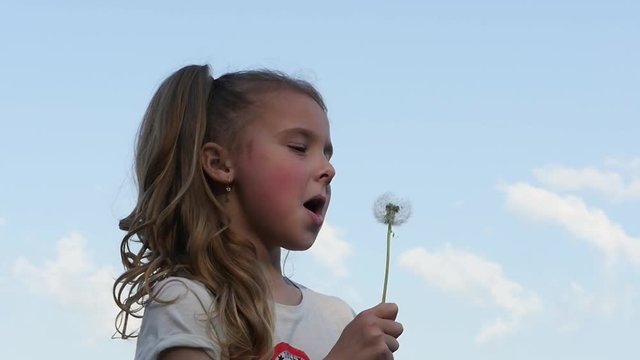 Lovely girl blowing dandelion on sky background repeatedly. Teenager girl looking into the camera turns to the side and blows a dandelion. Slow motion