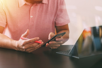 businessman in pink t-shirt working with smart phone and digitl tablet computer on wooden desk in modern office