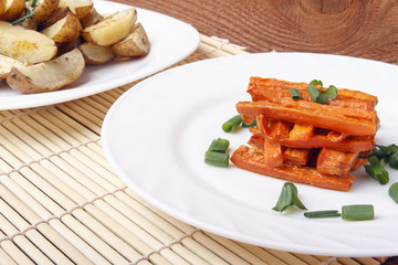 Baked carrots and potatoes with green onions on a white plate. Organic Vegetarian Food on a bamboo mat