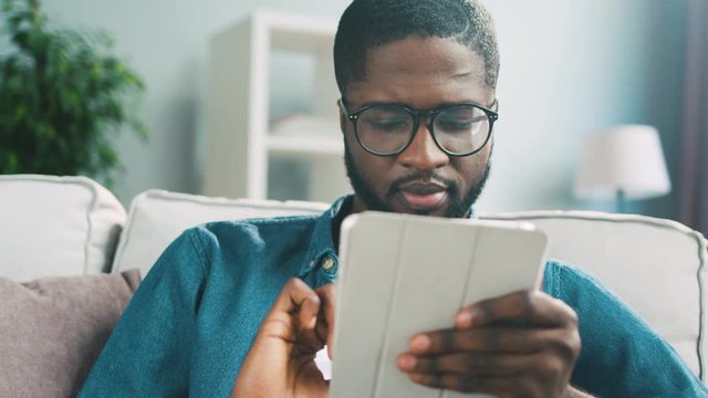 Young handsome african man in glasses sitting on sofa using laptop for shopping on living background. Close up.