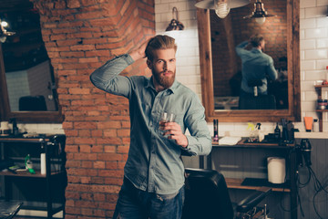 Casual men`s life. Chill out at the barber shop. Handsome young red bearded man in casual jeans outfit, drinking scotch and fixing his perfect haircut © deagreez
