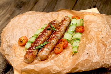 Fried sausages with cherry tomatoes, cucumber and rosemary on the parchment on the old wooden table.