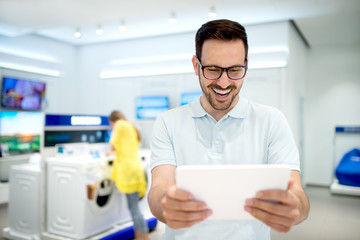 Fototapeta premium Smiling man playing a game on tablet in tech store. Buying new piece of technology.