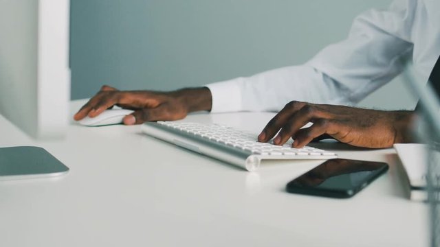 Side view of hands of african american man in office in white shirt typing using white computer keyboard on white table.