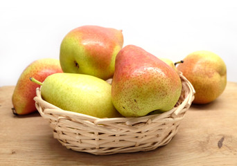 Red and yellow pears lays in small wicker straw basket on brown wooden table front view closeup