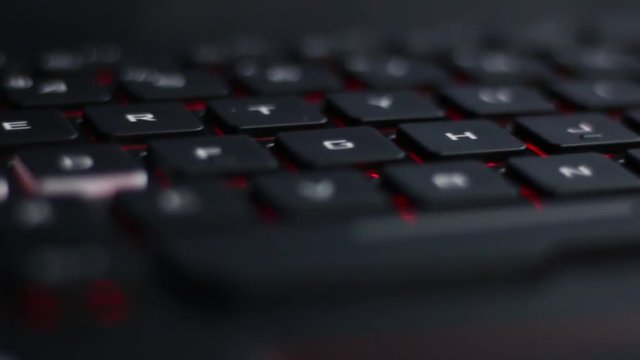 Angled Close up diagonal move left to right over length of  back lit laptop keyboard with red nahimic RGB keys shining from below 