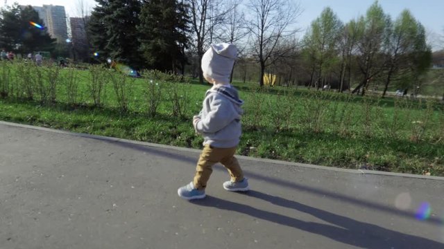 Little cute boy with confident rapid steps walks along the road in the park in summer at sunlight. In the background there are green lawns and trees. Beautiful summer day in the park