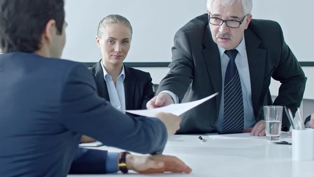 Senior manager giving document to new employee at job interview and discussing something with young female colleague while man signing application form