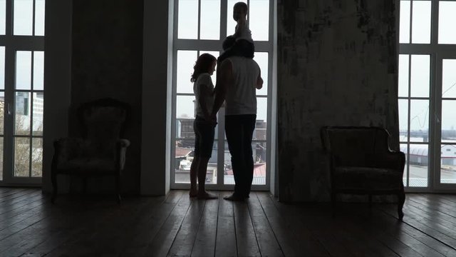 The young couple is by the window of the apartment. The young family approaches a window of the apartment. Siluyet of a young family at a window. Silhouette at the window.