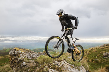 Obraz premium Ryder in full protective equipment on the mtb bike climbs on a rock against the backdrop of a mountain range and low clouds