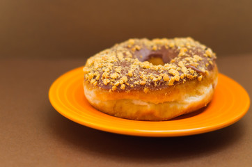 Delicious single donut with chocolate glazed with a powder peanut, over an orange plate in a soft brown background