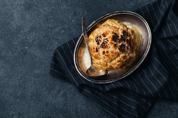 Whole roasted cauliflower with goat cheese sauce on metallic tray on dark rustic background with vintage spoon