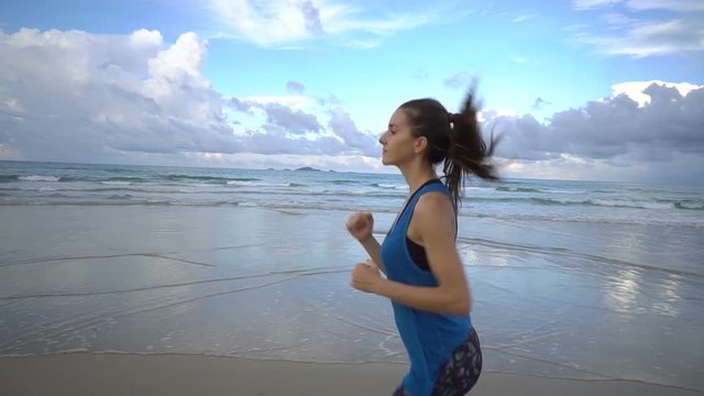 Healthy active woman in blue top running on the beach near sea on sunset.