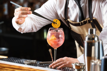 Elegant barman is making pink cocktail holding orange chips at bar counter background. - obrazy, fototapety, plakaty