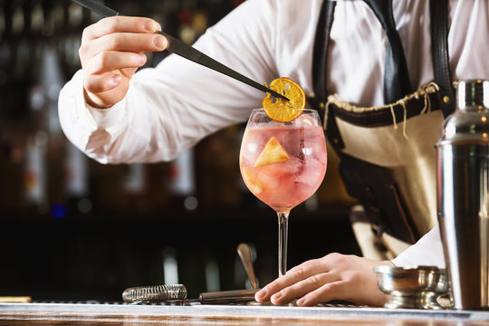 Elegant Bartender Is Preparing Pink Cocktail Holding Orange Chips At Bar Counter Background.