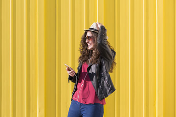 Portrait of a beautiful young woman listening to music on her mobile phone with earphones and having fun over yellow background. Casual clothing.  Wearing sunglasses and a modern hat. Happy.