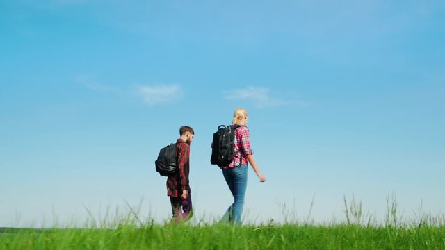 A man and a woman with backpacks are walking behind a green meadow against a background of a beautiful blue sky. Travel, tourism, a healthy life
