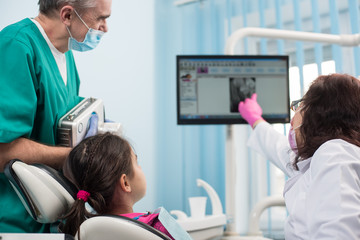 Senior pediatric dentist with assistant doing dental treatment patient girl using dental x-ray machine in dental office. People examine X-ray image on monitor. Focus on the girl. Dental equipment