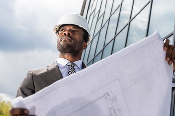 Low angle view of professional architect in hard hat holding blueprint outside modern building