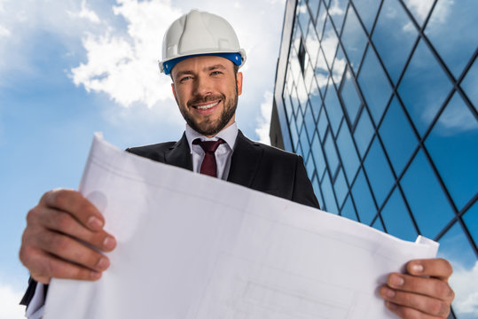 Low angle view of smiling bearded architect holding blueprint and looking at camera