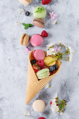 Variety of colorful french sweet dessert macaron macaroons with different fillings served in waffle cone with spring flowers and fresh berries over gray texture background. Top view with space