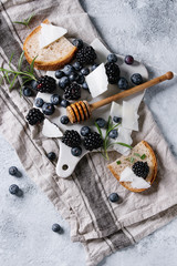 Fototapeta na wymiar Berries blackberry and blueberry, honey on dipper, rosemary, sliced goat cheese with bread served on ceramic board with textile linen over gray texture background. Summer sandwich. Top view with space
