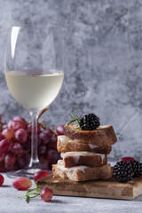 Grilled sandwich with melted goat cheese, blackberry, blueberry, rosemary and honey, served on wooden board with glass of white wine and grape over gray blue texture background. Summer appetizer.