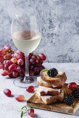 Grilled sandwich with melted goat cheese, blackberry, blueberry, rosemary and honey, served on wooden board with glass of white wine and grape over gray blue texture background. Summer appetizer.