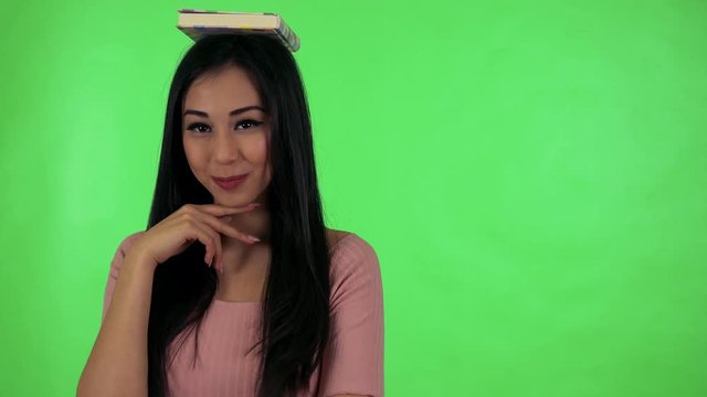 young attractive asian woman smiles to camera with book on head - green screen