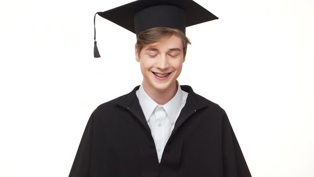 Young Caucasian graduate in black robe rearranging his square academic cap on white background