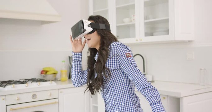 Beautiful young brunette in checkered shirt looking amazed while wearing goggles of virtual reality and standing in modern light kitchen.