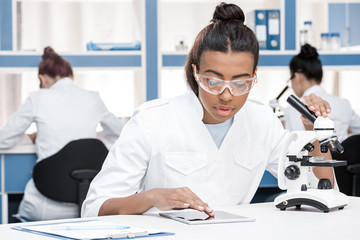 african american scientist in lab coat with microscope and digital tablet working in chemical lab, scientists group behind