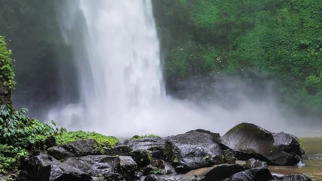 Close up of Nungnung waterfall, Falling water hitting water surface, some huge wet rocks in front of frame. Lush green leafes motion in background, Bali, Indonesia