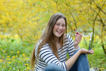 Beautiful young woman applying red lipstick looking at mirror isolated on natural background. Makeup and cosmetics concept.